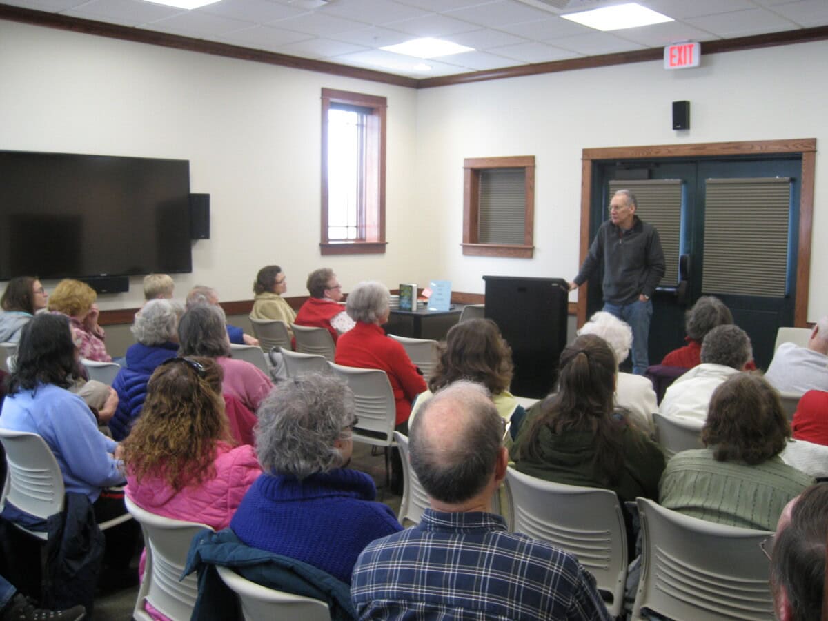 A man stands at a podium speaking to an audience seated in a small meeting room. Most attendees appear to be older adults, and they are listening attentively. A large TV and windows are visible in the background.