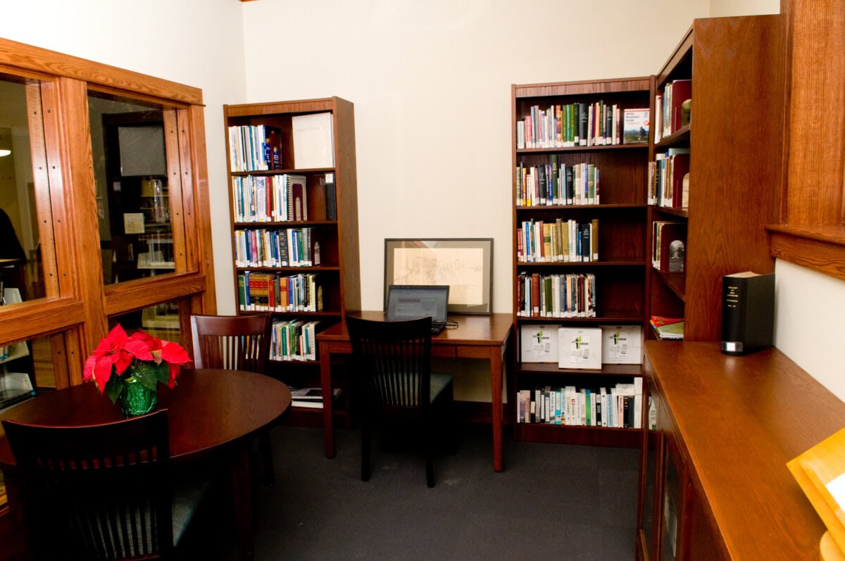 A small library room with wooden bookshelves filled with books, a desk with a chair and a laptop, a framed picture, a round table with chairs, and a red poinsettia plant on the table.