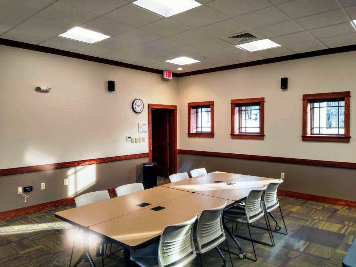 A conference room with beige walls, a rectangular table surrounded by white chairs, three small windows, a clock on the wall, and sunlight streaming in through the windows.