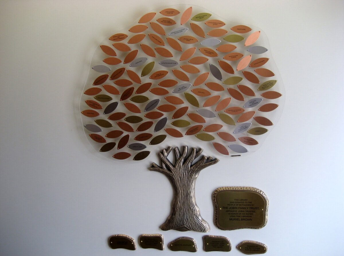 A decorative metal tree mounted on a wall, with copper, gold, and silver leaves engraved with names. Below the tree trunk are plaques with additional inscriptions.