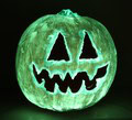 A glowing green jack-o’-lantern with triangular eyes and a jagged, toothy smile is lit up against a dark background.