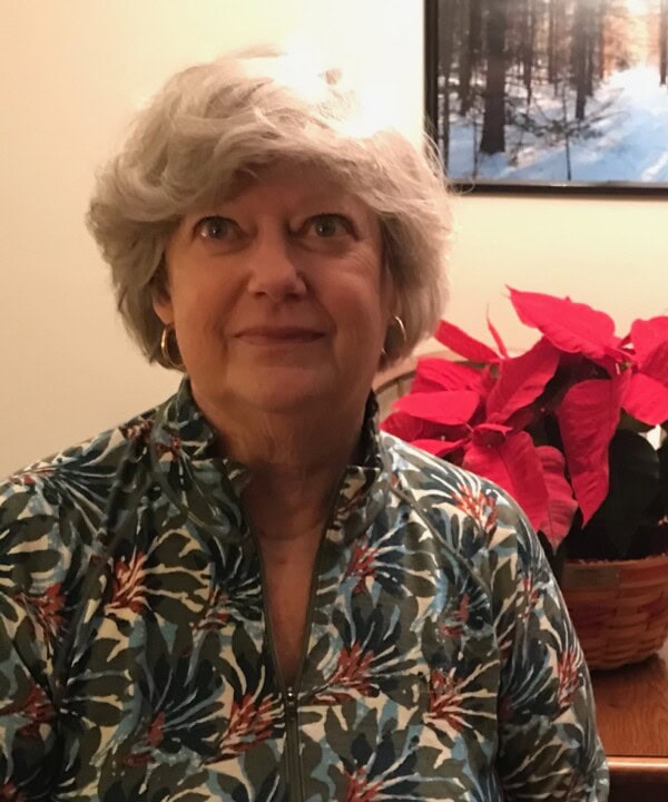 An older woman with short gray hair and gold hoop earrings smiles slightly, wearing a patterned jacket. She is indoors near a red poinsettia plant and a framed picture of a snowy forest on the wall behind her.