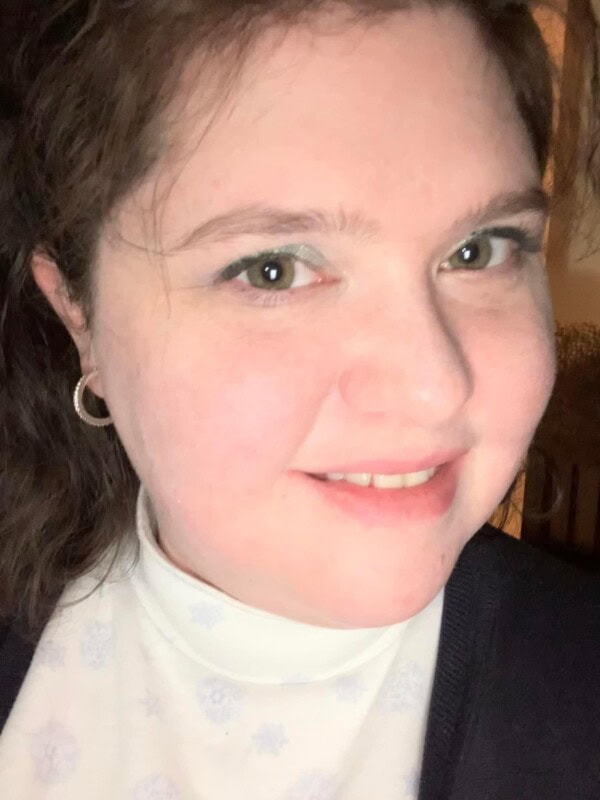 A woman with fair skin and dark brown hair smiles at the camera, wearing a white turtleneck with a subtle pattern, a dark sweater, and small hoop earrings. The background is softly lit and out of focus.