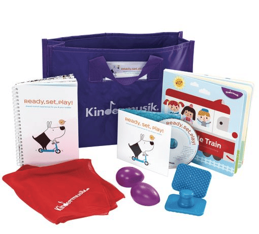 A purple Kindermusik tote bag with a spiral notebook, a red scarf, a book, a CD, two purple egg shakers, and a blue textured block displayed in front of it.