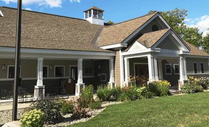 A single-story building with a gabled roof, covered front porch, white columns, and landscaped garden beds, set against a blue sky with scattered clouds.