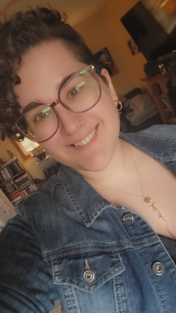A person with short, curly hair and glasses smiles at the camera. They are wearing a denim jacket and a necklace, with a cozy, well-lit room and bookshelves in the background.