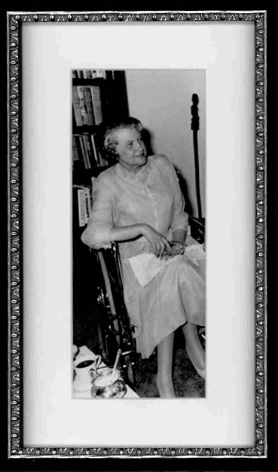 Black-and-white photo of an older woman sitting in a wheelchair, wearing a light-colored dress, holding papers, and looking slightly upward. Framed with a decorative border; bookshelves and a table are visible in the background.