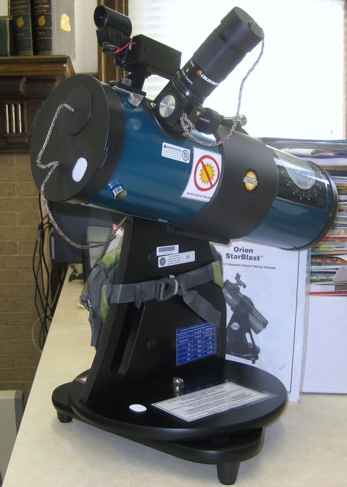 A blue Orion StarBlast tabletop telescope sits on a black mount, with a finder scope and eyepiece attached. The telescope is indoors, next to its instruction manual and a stack of papers.