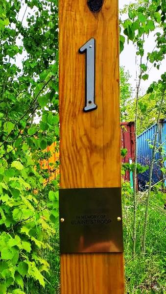 A wooden post with a metal number 1 and a small metal plaque reading In memory of Clarence Stroop, surrounded by green foliage and colorful shipping containers in the background.